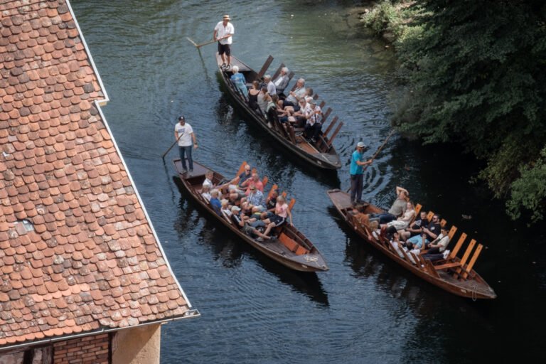 Groups of people riding in Stocherkähne down the Neckar. They sit on wooden boards. Stocherer stand at the end with long wooden poles.
